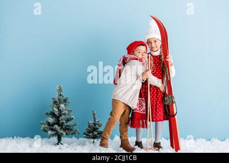 boy in hat and scarf hugging sister with ski poles and skis while standing on blue Stock Photo