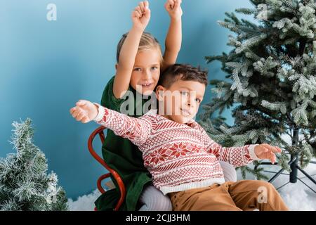 excited children with hands above heads sitting in sleigh near green pines on blue Stock Photo