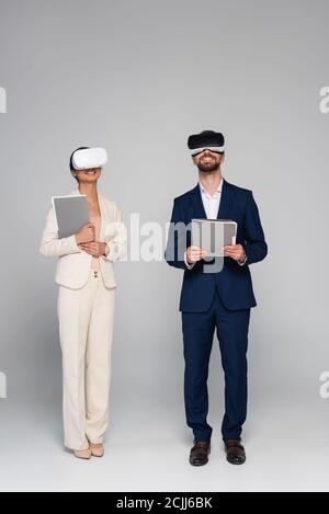 full length view of businesspeople in suits and vr headsets holding documents while standing on grey Stock Photo
