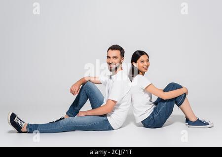 young asian woman and bearded man in white t-shirts and jeans sitting on white and looking at camera Stock Photo