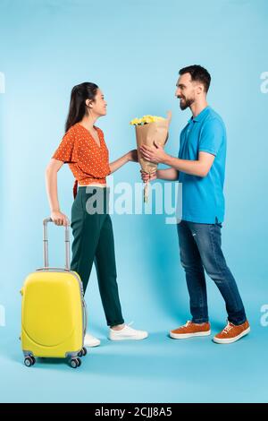 man in polo t-shirt and jeans presenting bouquet to asian woman with yellow suitcase on blue Stock Photo