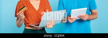 cropped view of woman in red blouse holding credit card and laptop near man with digital tablet on blue, panoramic concept Stock Photo