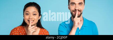 panoramic concept of asian woman in red blouse and man in polo t-shirt showing silence gesture isolated on blue Stock Photo