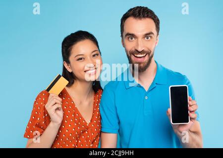 bearded man in polo t-shirt showing smartphone with blank screen near asian woman holding credit card isolated on blue Stock Photo