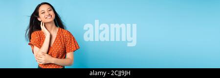 horizontal concept of elegant asian woman in red blouse looking at camera while touching neck on blue Stock Photo