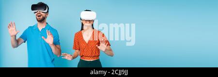 panoramic concept of excited man in polo t-shirt and woman in red blouse gesturing with hands while using vr headsets on blue Stock Photo