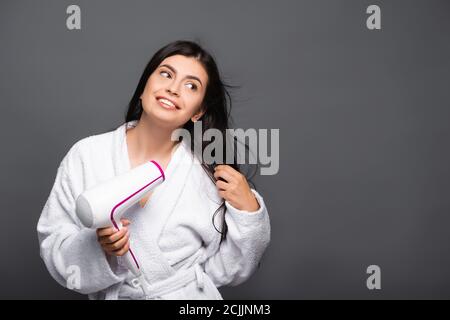 brunette long haired woman in bathrobe using hairdryer isolated on black Stock Photo