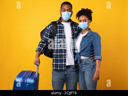 African Tourists Wearing Protective Masks Standing With Suitcase, Yellow Background Stock Photo