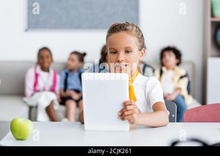 Selective focus of schoolgirl using digital tablet near apple and multicultural friends at background in classroom Stock Photo