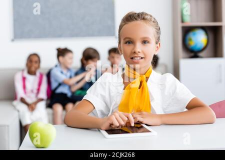 Selective focus of schoolgirl using digital tablet near apple and multicultural classmates in classroom Stock Photo