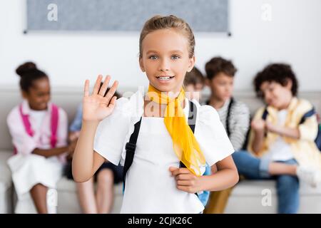 Selective focus of schoolgirl with backpack waving hand at camera with multicultural friends at background Stock Photo