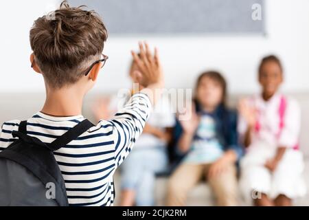 Selective focus of schoolboy with backpack waving hand at multiethnic classmates Stock Photo