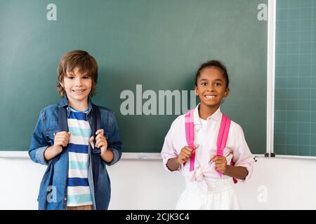 Schoolboy with backpack standing near african american friend and chalkboard in classroom Stock Photo