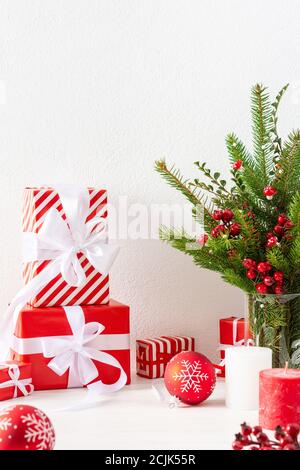 Gift boxes and red balls with candles, fir tree branches and hawthorn twigs in a vase on white wall background, new year and christmas concept Stock Photo