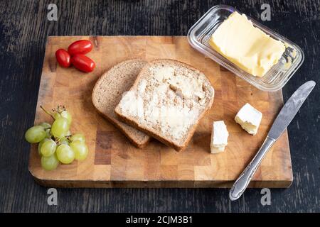 Lunchtime sandwich making – two slices of bread, butter, cheese, tomatoes and grapes, on a wooden cutting board Stock Photo