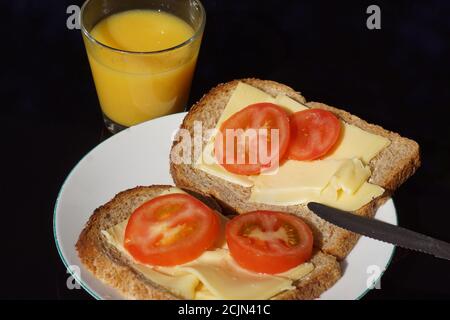 White plate with two slices of bread topped with cheese and tomato on a black glass table top. A serrated knife and a glass of orange juice. September Stock Photo