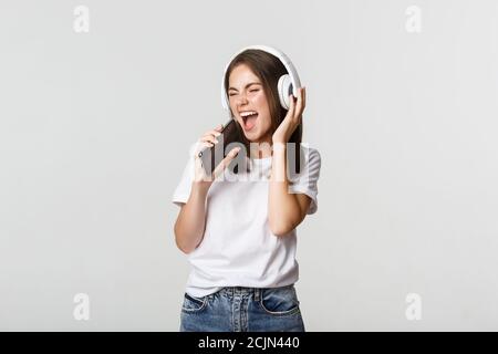 Attractive happy woman playing karaoke app, singing into smartphone, wearing wireless headphones Stock Photo