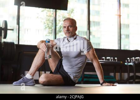 Portrait of mature man sitting on the floor and drinking water from the bottle he resting after sports training in gym Stock Photo
