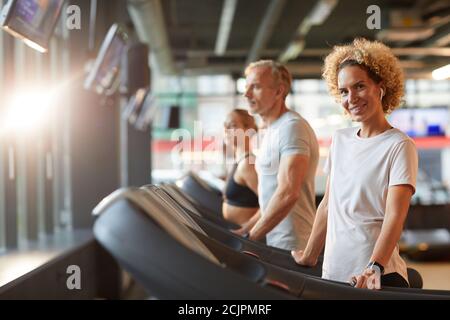 Portrait mature woman smiling at camera during her training on treadmill with other people in the background Stock Photo