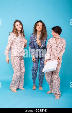 Portrait of three beautiful young girls wearing colorful pyjamas having fun during sleepover isolated over blue background. Pajama party and hen-party Stock Photo