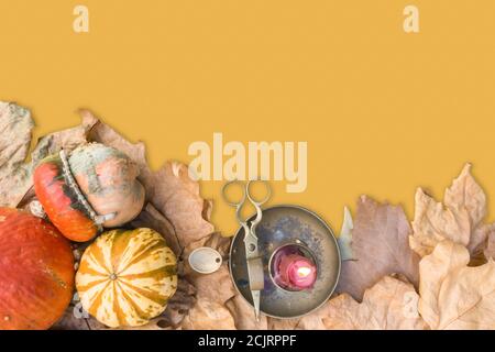 Various raw pumpkins with dry fall leaves and burning candle on old bronze candlestick with scissors yellow background. Halloween vintage background. Stock Photo
