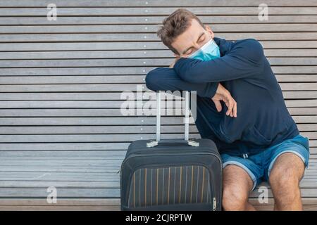 Man in disposble medical mask sleeping on bench leaning on luggage. Traveler wearing protective face mask with suitcase waiting for trip. New normal Stock Photo