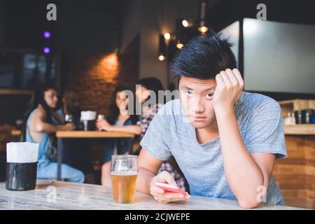 A sad handsome excluded asian boy sitting alone in a bar using his smartphone Stock Photo