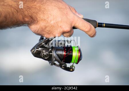 Close-up fisherman's hands reeling in the reel at the seashore. The fisherman's hands, holds the spinning rod, rotates the coil handle. Hobby and Stock Photo