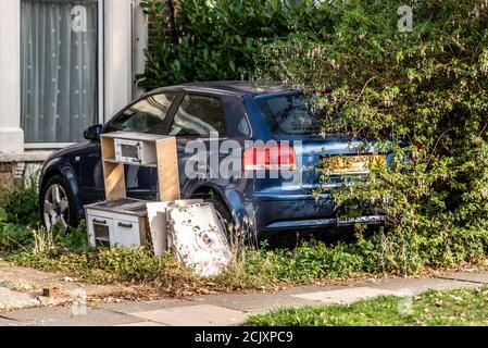 Overgrown car in front garden of house, with rubbish. Dumped kitchen cabinets outside in front yard, blocking car. Blue Audi A3 left to decay in bush Stock Photo