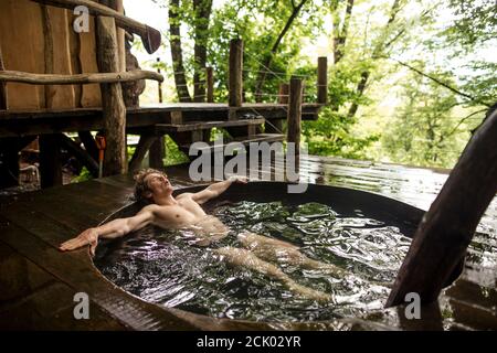 pleasant young man with closed eyes is enjoying taking bath in beautiful landscape near the wooden house. spa treatment, health care concept Stock Photo
