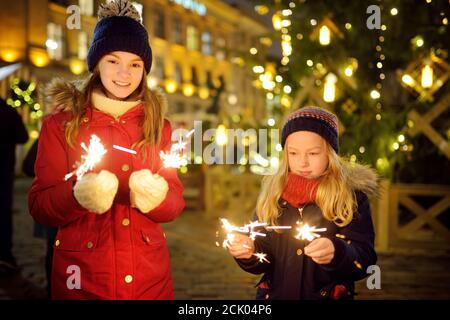 Two adorable sisters holding sparklers near a Christmas tree on traditional Christmas fair in Riga, Latvia. Kids celebrating New Years eve. Winter tim Stock Photo