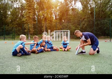 Young coach with clipboard teaches kids strategy of playing on football field. Stock Photo