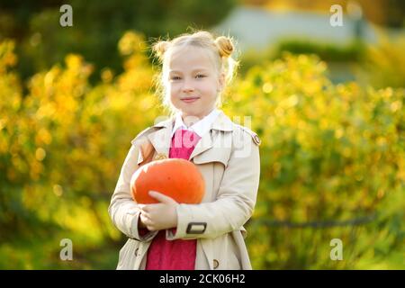 Cute young girl holding small pumpkin on a pumpkin patch. Kid picking pumpkins at country farm on warm autumn day. Family time at Thanksgiving and Hal Stock Photo
