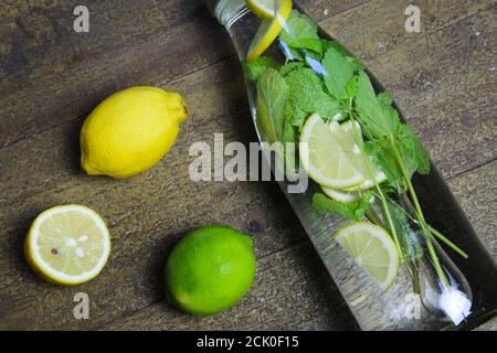 View on isolated water glass bottle flavored with mint leaves and yellow citrus, green lime fruit on wood table (focus on bottle) Stock Photo