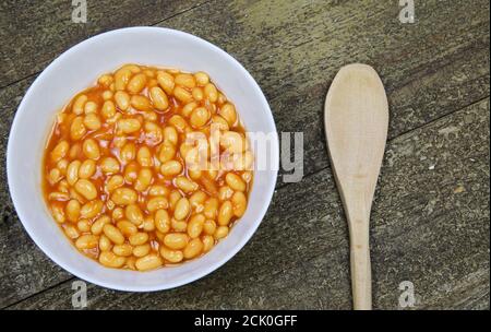 High angle view on isolated white bowl with baked beans in red sauce and wood spooon on wooden table Stock Photo