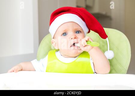 a baby in a Santa Claus hat is sitting in a high chair and eating with a spoon Stock Photo