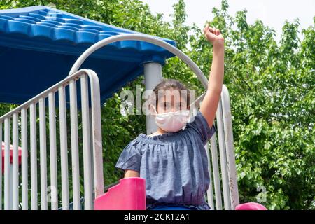 Little girl at playground surrounded by trees, wearing a protective face mask and holding her arm up to protest the Covid-19 quarantine Stock Photo