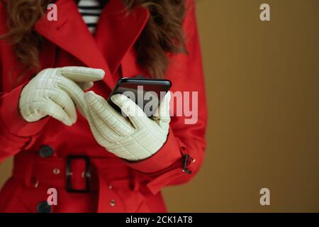 Life during covid-19 pandemic. Closeup on woman in red coat sending text message using smartphone against bronze background. Stock Photo