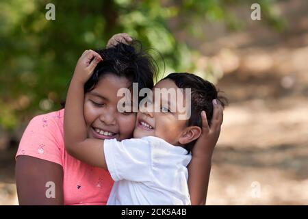 Little brother is hugging his older sister with his arms around her face and both are smiling in outdoor setting.