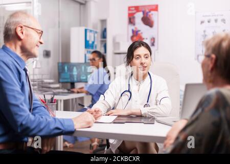 Young woman doctor examining old couple in hospital office wearing white coat and stethoscope. Pensioner pair on medical check and nurse working on computer. Stock Photo