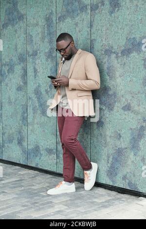African young businessman leaning on the wall of building and typing a message on his mobile phone Stock Photo