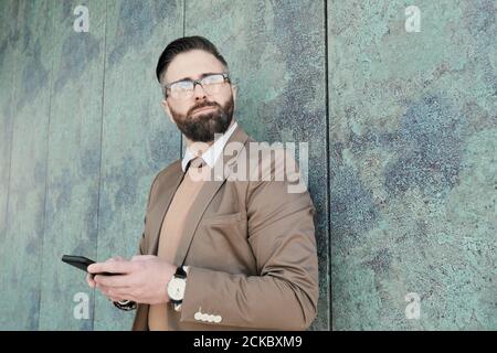 Bearded confident businessman in eyeglasses using his mobile phone while standing outdoors Stock Photo