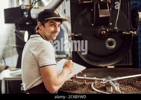 Joyful male worker taking notes while using coffee roasting machine Stock Photo