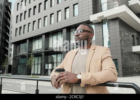 Young African man in eyeglasses drinking coffee while standing outdoors he enjoying the view of the city Stock Photo