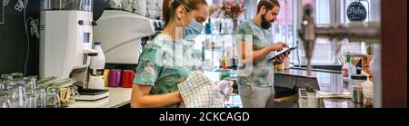 Waitress with mask cleaning glasses while her coworker works wit Stock Photo