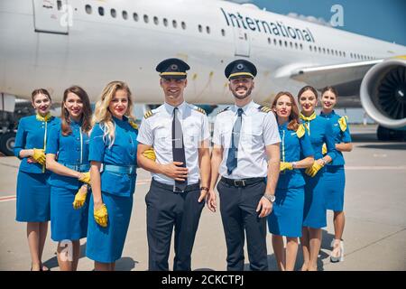 Handsome pilots and beautiful stewardesses looking at camera and smiling while posing at airport with airplane on background Stock Photo