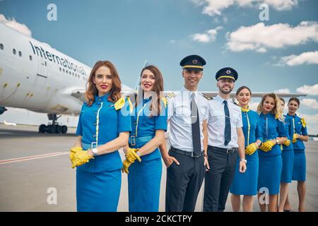 Beautiful young stewardesses and pilots looking at camera and smiling while posing at airport with airplane on background Stock Photo