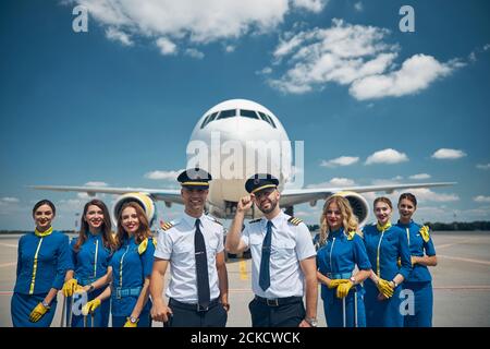 Beautiful stewardesses and handsome pilots looking at camera and smiling while posing at airport with airplane on background Stock Photo