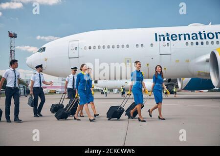 Cheerful airline workers in uniforms carrying trolley luggage bags while walking down the airfield Stock Photo