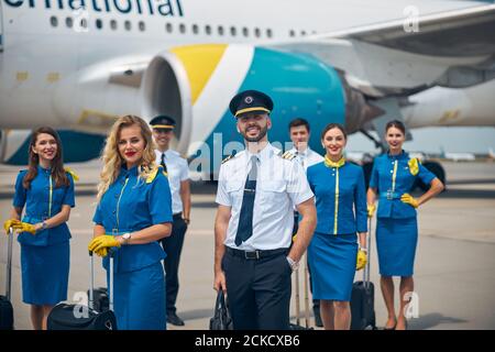 Charming stewardesses and handsome pilots in uniforms looking at camera and smiling while posing at airport with airplane on background Stock Photo